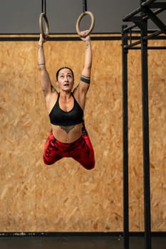 Vertical photo of a sportive woman hanging from olympic rings while working out in a gym