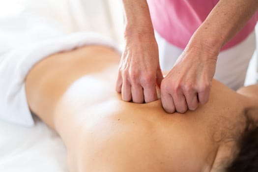 High angle of anonymous osteopath using fist while massaging back bone of unrecognizable topless female patient lying on bed during rehabilitation session in modern clinic