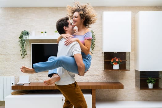 Side view of cheerful young couple in casual clothes smiling and hugging while having fun at home