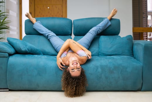 Happy young female in jeans and top with curly hair lying upside down on sofa in living room and having fun at home while enjoying