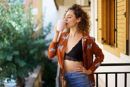 Young stylish female with curly brown hair wearing casual clothes drinking coffee while standing on balcony of modern apartment in town against green background