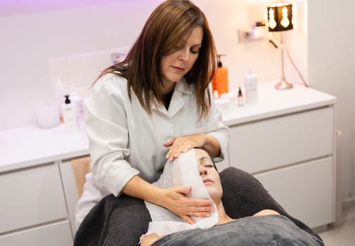 Professional female beautician looking down, while standing near beauty salon bed with anonymous client with closed eyes and treating face with hot water wet towel in lights