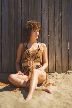Full body of barefoot female in swimwear and knitted beach dress sitting on sand leaning on wooden fence looking away thoughtfully during sunny day