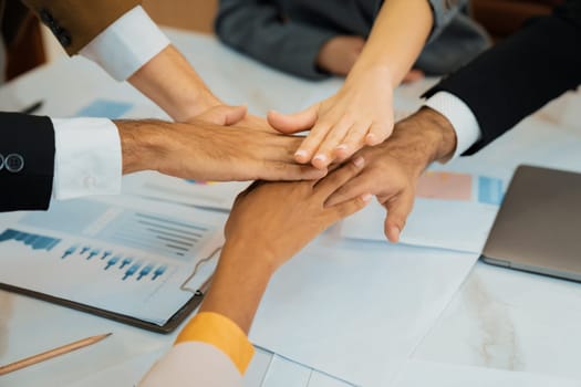 Top view of corporate diverse businesspeople putting hands together on meeting table with document scatter around at business meeting room. Represented unity, cooperation, collaboration. Ornamented.