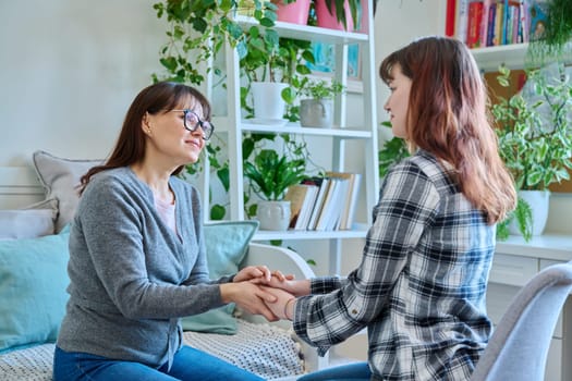 Talking mother and teenage daughter sitting together on couch at home, smiling holding hands. Family, communication, motherhood, friendship, relationship between parent and daughter 18-20 years old