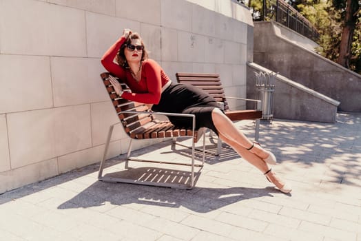 Portrait of a woman on the street. An attractive woman in glasses, a red blouse and a black skirt is sitting on a bench outside