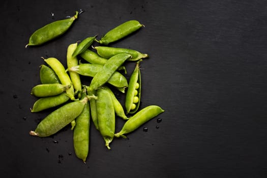 Fresh green pea pods scattered on a dark slate background, some opened showing peas inside.