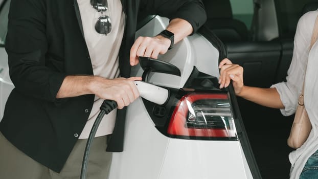 Young couple travel with EV electric car to shopping center parking lot charging in downtown city showing urban sustainability lifestyle by green clean rechargeable energy of electric vehicle innards