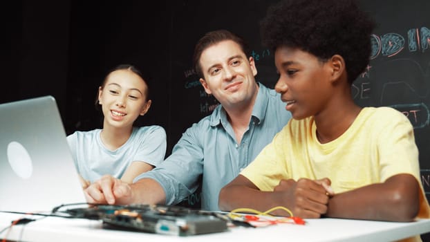 Caucasian teacher talking about electric tool while student fixing robotic model on table with electronic equipment placed. Diverse academic children learning about main board structure. Edification.