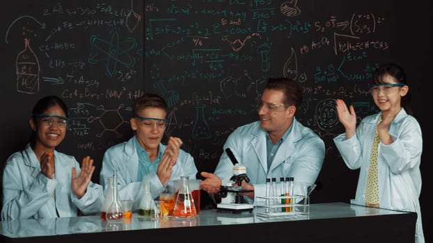 Diverse student clap hands while teacher show thumb to celebrate successful experiment. Edification.. Caucasian teacher looking after highschool children doing experiment at blackboard. Edification.