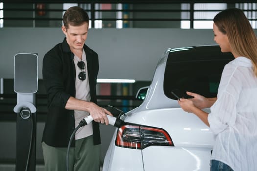 Young couple travel with EV electric car to shopping center parking lot charging in downtown city showing urban sustainability lifestyle by green clean rechargeable energy of electric vehicle innards