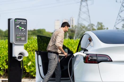 Young man recharge EV car battery at charging station connected to power grid tower electrical industrial facility as electrical industry for eco friendly vehicle utilization. Expedient