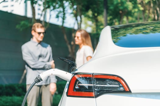 Young couple travel with EV electric car charging in green sustainable city outdoor garden in summer shows urban sustainability lifestyle by green clean rechargeable energy of electric vehicle innards