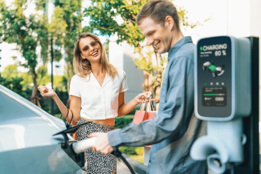 Young couple travel with EV electric car charging in green sustainable city outdoor garden in summer shows urban sustainability lifestyle by green clean rechargeable energy of electric vehicle innards