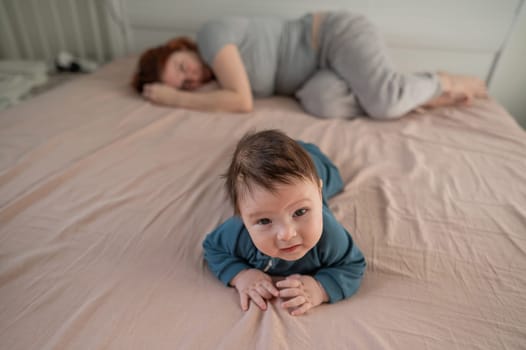 A three-month-old boy lies on his stomach on the bed and his mother sleeps behind him. Postpartum depression