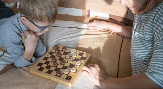 A caring middle-aged grandmother plays checkers with a child, a boy, lying on the bed. Happy family enjoying an interesting wooden board game at home