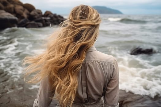 Rear view of a blonde girl on the shore of a stormy sea. Photo from the back of a woman looking over the horizon of the sea.