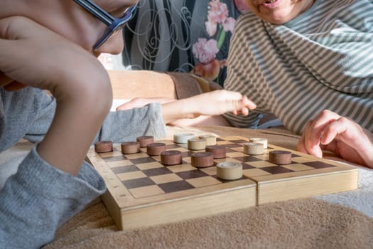 Close-up of a caring middle-aged grandmother playing checkers with a child, a boy, while lying on the bed. Happy family enjoying an interesting wooden board game at home