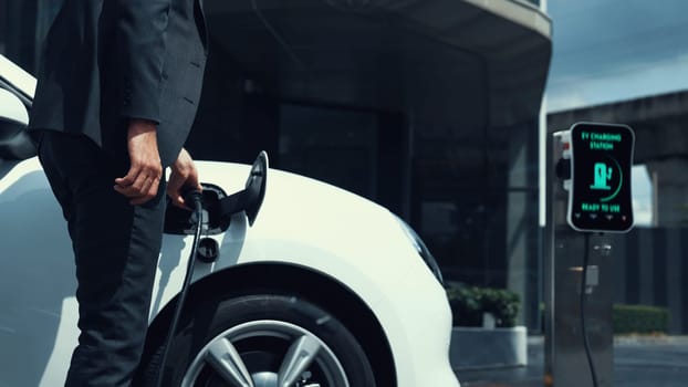 Businessman recharge his electric car from charging station at city center or public car park. Eco friendly rechargeable car using alternative clean energy in urban city lifestyle.Peruse