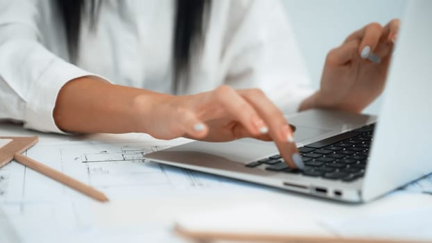 Close-up of young beautiful professional architect hands using laptop with blueprint and architectural document placed on table at modern office. Creative design concept. Focus on hand. Immaculate.