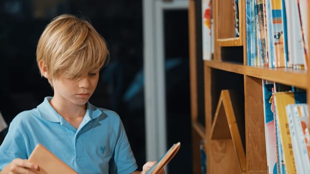Young smart caucasian boy picking or choosing from book shelf and reading a book. Clever child learning, studying, open a books at library. Attractive kid turning page. Stack of books. Erudition.