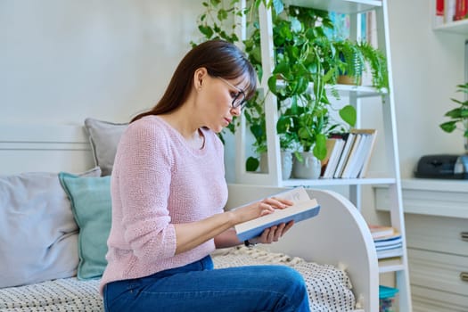 Mature woman reading paper book sitting on couch at home. Relaxation, literature hobby, fiction, lifestyle, people concept