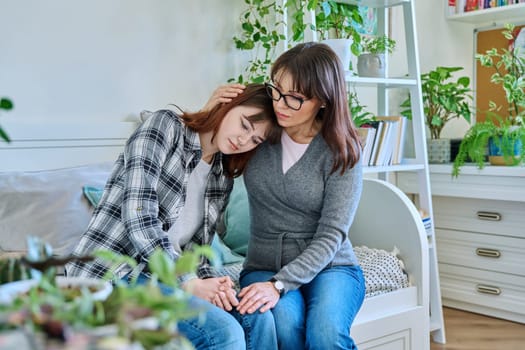 Upset sad hugged mother and daughter teenager sitting together on the couch at home. Family, communication, motherhood, friendship, relationship between parent and teen daughter concept