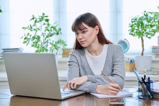 College student girl sitting at desk using laptop computer, making notes in study notebook, at home. Teenager female watching webinar, preparing for exam tests, studying remotely.