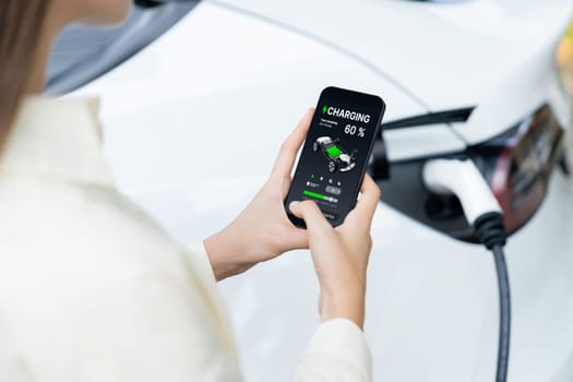 Young woman use smartphone to pay for electricity at public EV car charging station green city park. Modern environmental and sustainable urban lifestyle with EV vehicle. Expedient