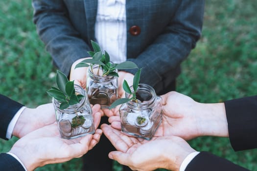 Concept of sustainable money growth investment with glass jar filled with money savings coins with businesspeople as eco-friendly financial investment nurtured with nature and healthy retirement. Gyre