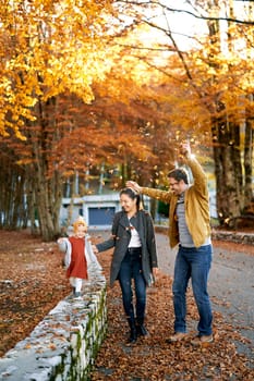 Mom leads a little girl by the hand along the fence in the park next to dad sprinkling leaves on them. High quality photo