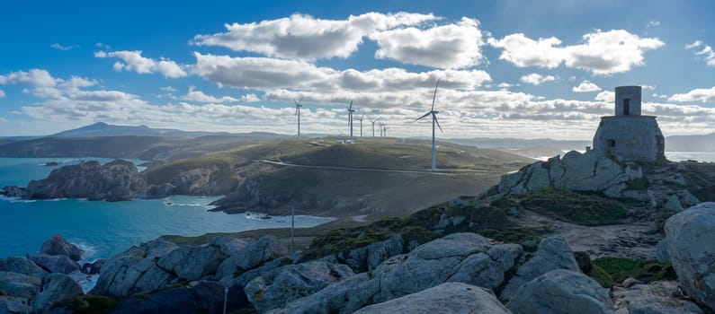 An ancient stone tower stands on a rocky outcrop, a silent testament to the past amid the modernity of wind turbines crowning the rolling hills. This contrast is set against a dynamic sky, where the force of wind is both harnessed by technology and embodied in the wild sea below.