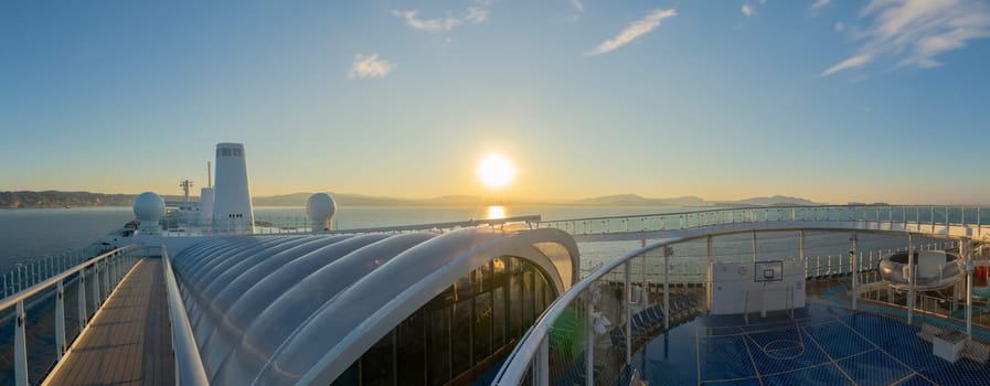 The golden sunrise casts a warm glow over a cruise ship deck, with sleek architectural lines leading towards the sun-kissed horizon. The peaceful ocean and distant mountains set a serene scene, inviting passengers to a day of maritime adventure.