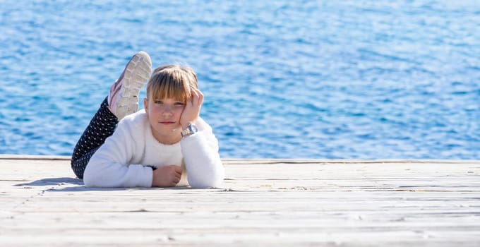 A young person reclines on a wooden pier, lost in thought, with the vast blue sea as their backdrop. The image captures a moment of youth and reflection, underscored by the infinite horizon and the tranquil maritime setting