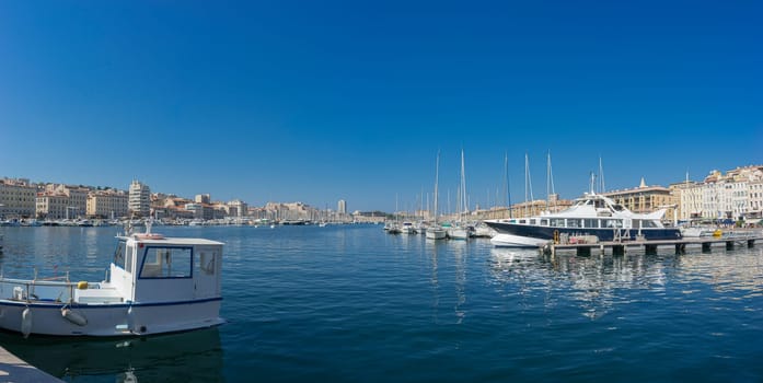 A clear day at the bustling port of Marseille, where a variety of boats and yachts are moored in calm blue waters. The city's historic architecture forms a charming backdrop, reflecting its rich seafaring heritage under the bright Mediterranean sun