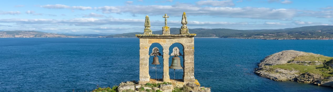 An ancient bell tower rises as a solitary sentinel on the edge of a cliff, its weathered stones and aged bells facing the endless blue sea. This historic watchtower offers a commanding view of the rugged coastline and distant hills, under the expansive sky