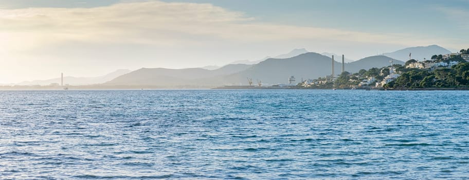 A coastal landscape where industry meets nature; factories and cranes are seen in the distance, harmonizing with the rolling mountains and the calm sea under a soft sky