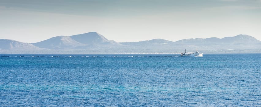 A fishing trawler cuts through the cobalt blue waters, its wake creating a frothy trail, set against the serene backdrop of distant mountains under a clear sky.