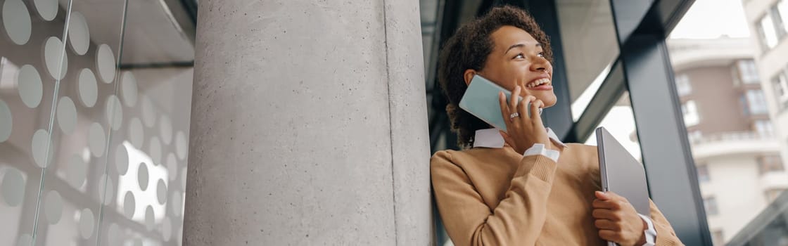 Stylish business woman is talking phone while standing with laptop in modern office