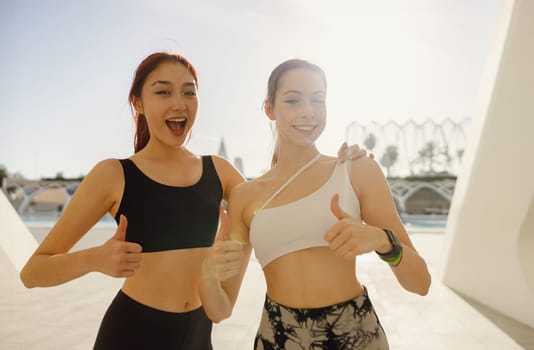 Two young female sportswomen have a rest after morning jogging outdoors and looks camera with smile