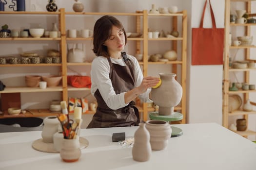 Smiling young female ceramist wearing apron work with unfired clay vase in pottery studio