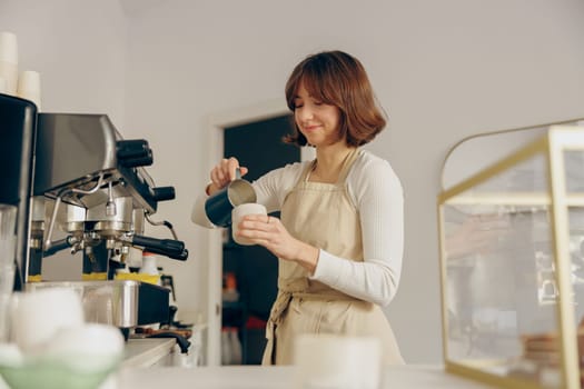 Woman barista in apron pouring hot milk foam into coffee. Professional coffee brewing concept