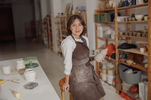 Portrait of young female potter in apron with mug looking at camera while posing in workshop