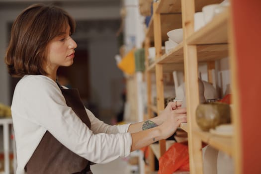 Female ceramist standing near shelves of clay mugs working in pottery studio. High quality photo