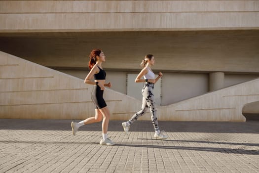 Two active women athlete running side by side along an outdoor track on modern buildings background