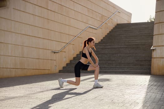 Young sportswoman in tracksuit stretches legs outside on modern building background