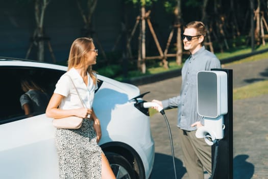 Young couple travel with EV electric car charging in green sustainable city outdoor garden in summer shows urban sustainability lifestyle by green clean rechargeable energy of electric vehicle innards