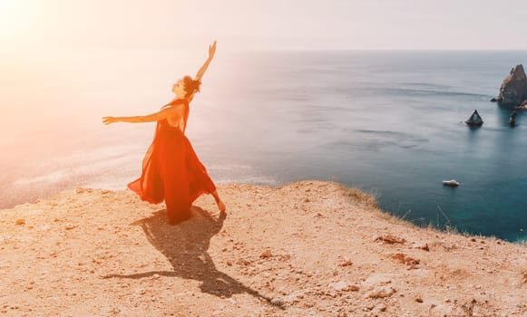 Side view a Young beautiful sensual woman in a red long dress posing on a rock high above the sea during sunrise. Girl on the nature on blue sky background. Fashion photo.