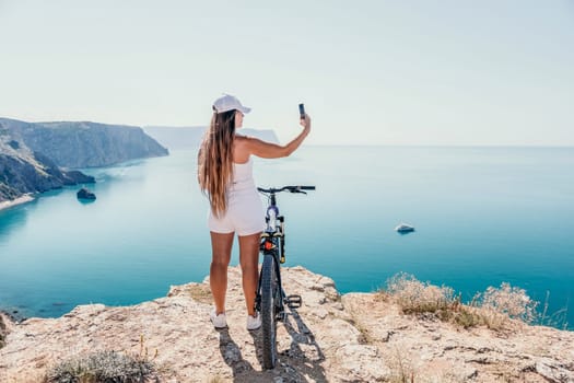 A woman cyclist on a mountain bike looking at the landscape of mountains and sea. Rear view of cyclist woman standing in front to the sea with outstretched arms. Freedom and healthy lifestyle concept