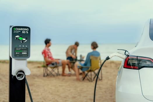 Alternative family vacation trip traveling by the beach with electric car recharging battery from EV charging station with blurred family enjoying the seascape background. Perpetual